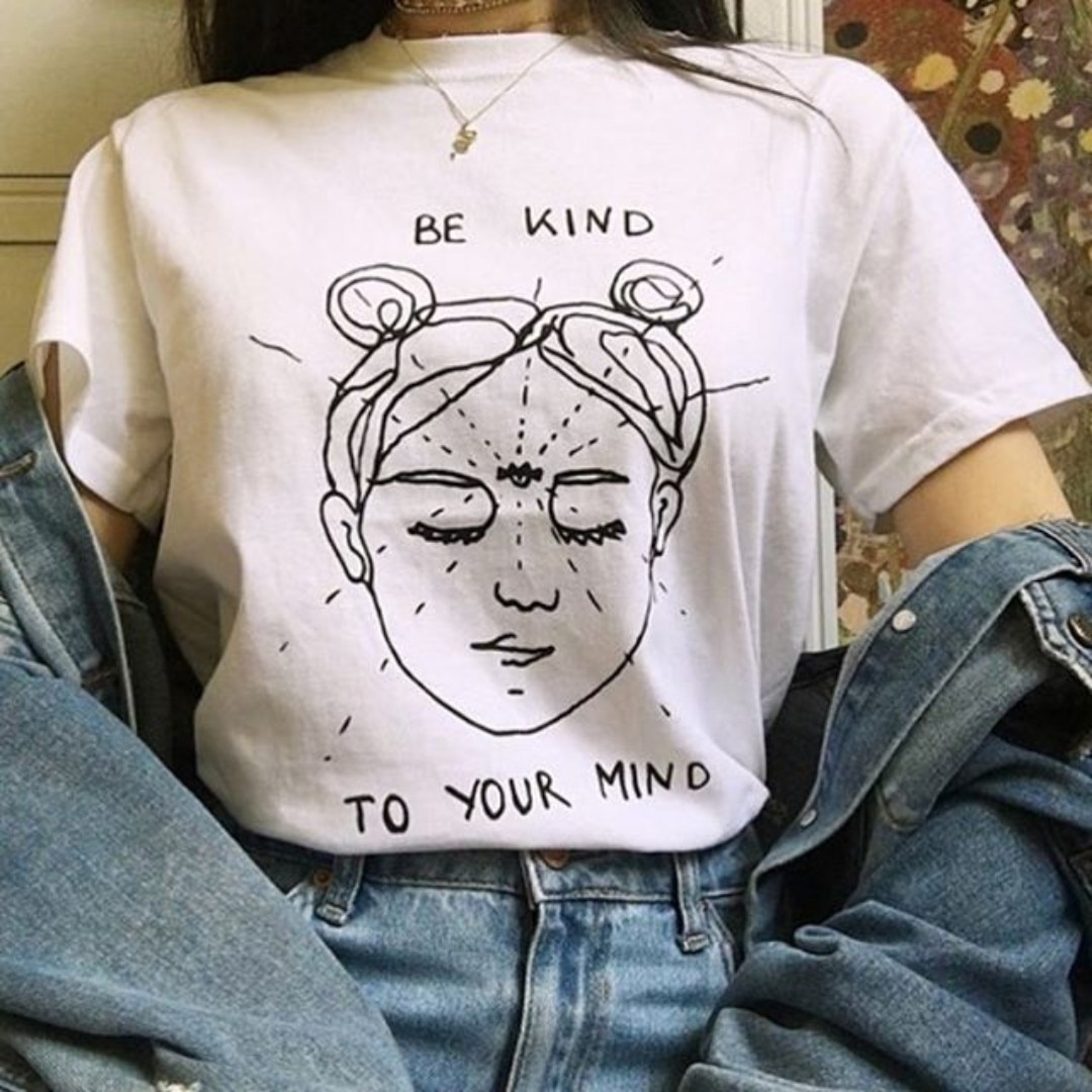 Woman wearing a white t-shirt with "Be Kind to Your Mind" graphic and line drawing of a peaceful face, paired with high-waisted jeans and a denim jacket.