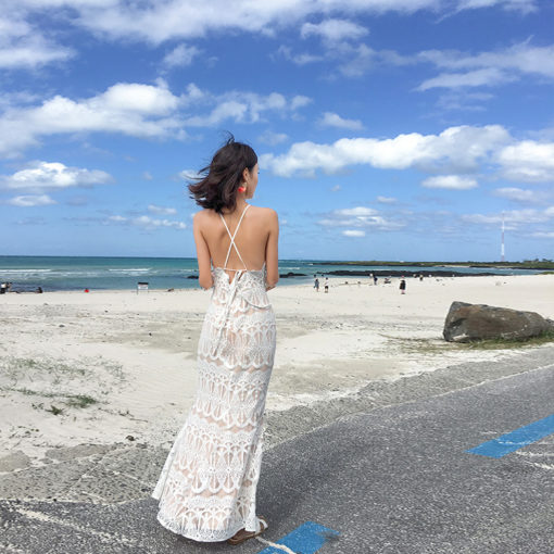 Woman wearing a white crochet maxi dress with a halter neckline, walking along a stone wall in a beach setting, showcasing an elegant and bohemian look.